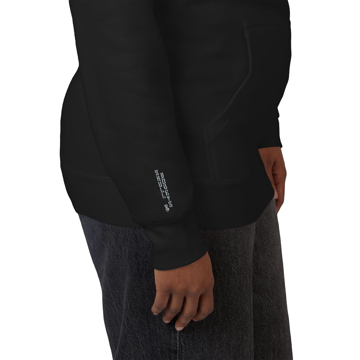 Close-up side view of a person wearing a black long-sleeve hoodie with a small white vertical logo on the cuff, paired with dark gray textured pants, against a plain white background.
