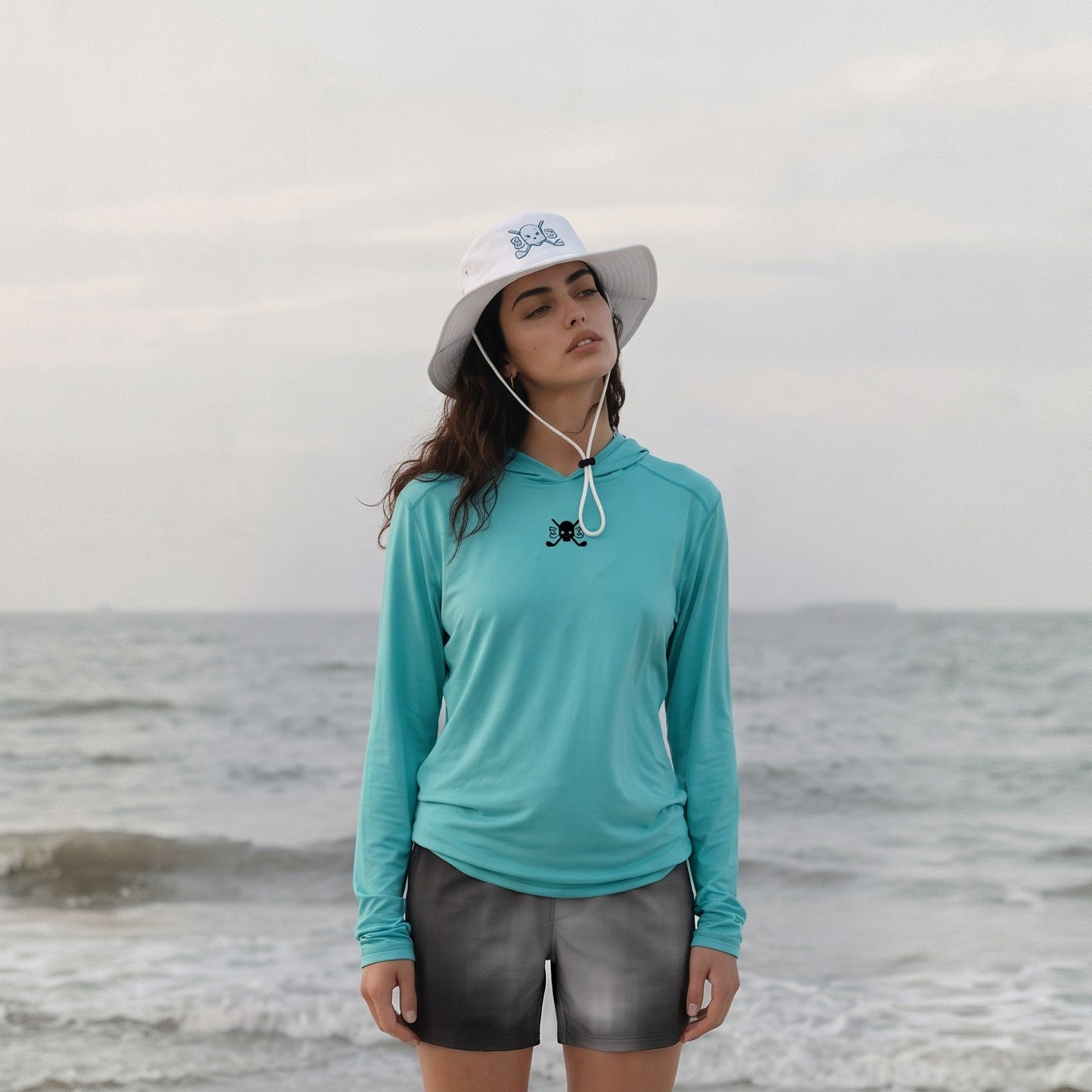 A woman stands on a white folding chair on a sandy beach, wearing a turquoise long-sleeve athletic hoodie, dark gray shorts, and a white sun hat with a blue logo. The ocean and overcast sky are in the background.
