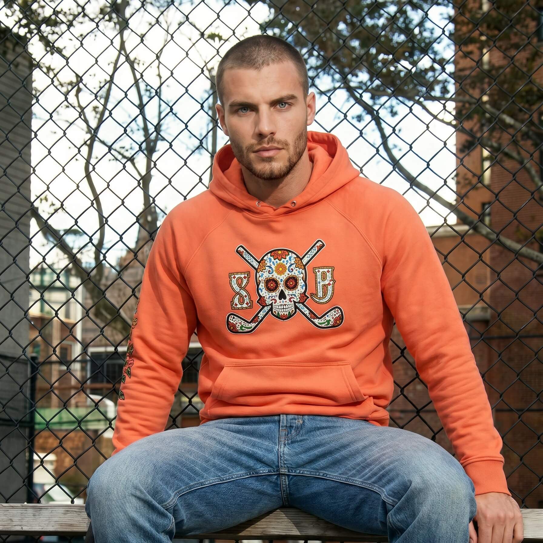 Man wearing an orange hoodie with a skull design, sitting on a bench outdoors.
