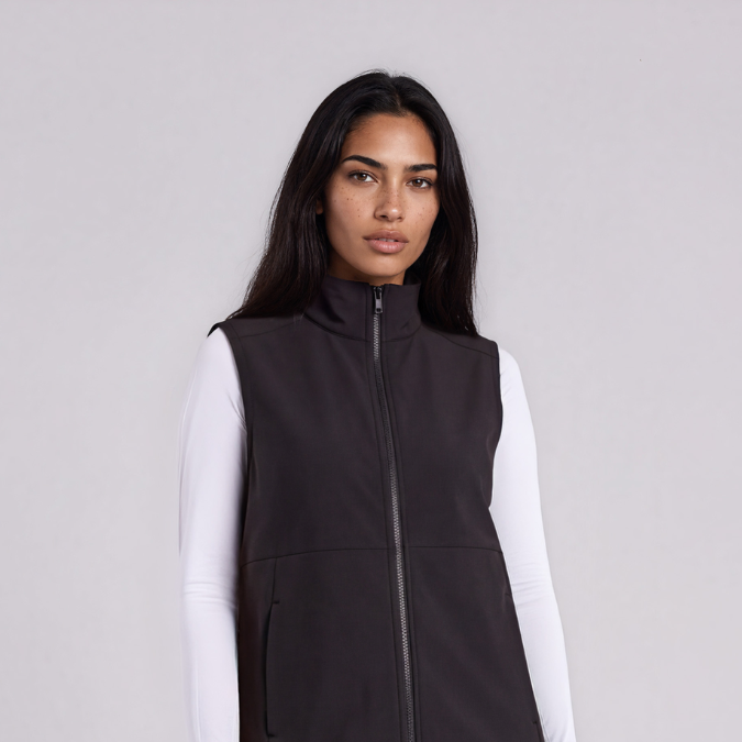 A woman modeling a black zip-up sleeveless vest over a white long-sleeve top, paired with dark gray skinny jeans and black ankle boots, against a light gray studio background.