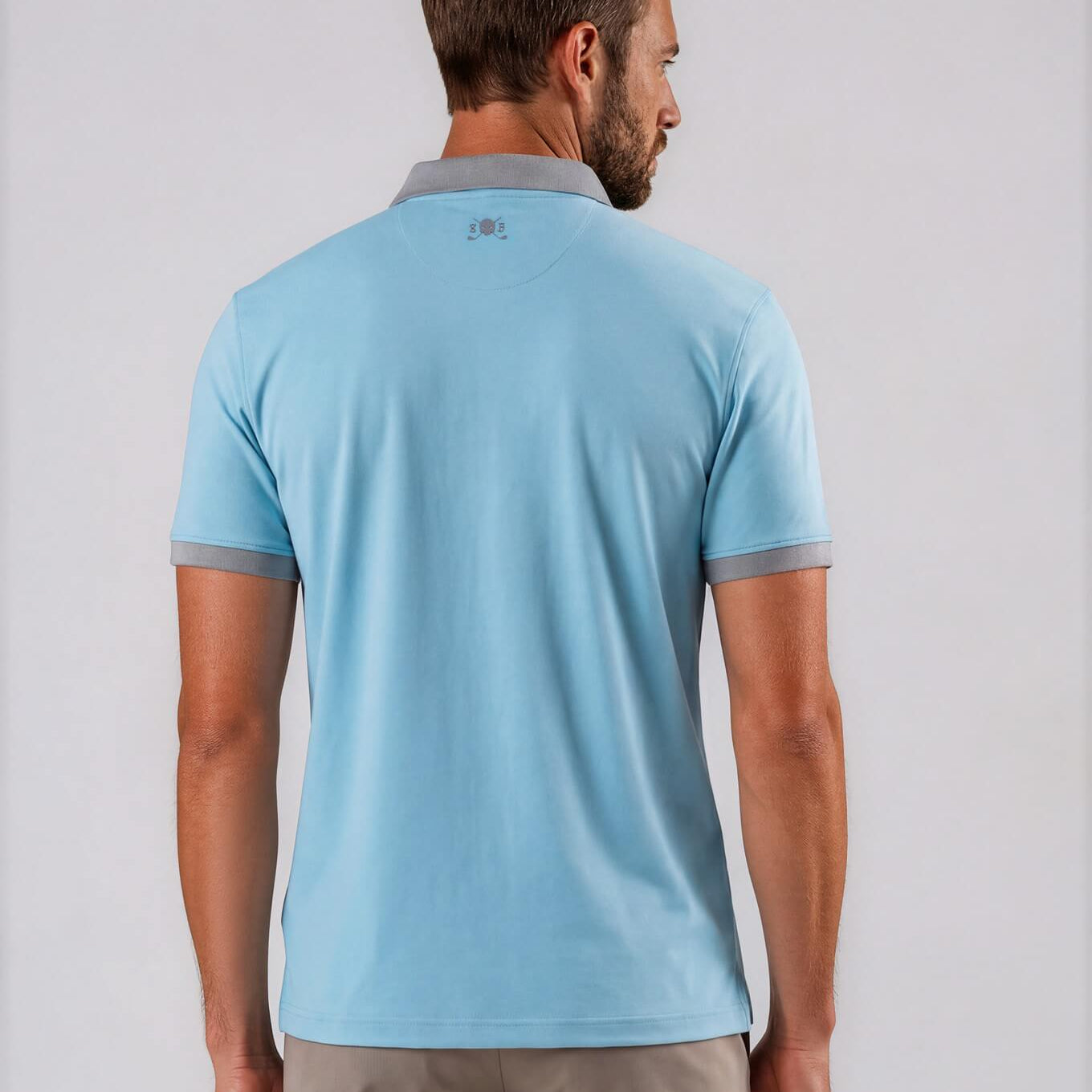 A man viewed from the back wearing a light blue Skull & Bogeys short-sleeve polo shirt with gray trim on the collar and sleeve cuffs, paired with beige trousers, against a light gray studio background.