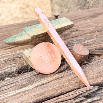 A copper-colored metal pencil with a pointed tip and a flat end rests on a rustic wooden surface next to a copper penny and a small metal object.
