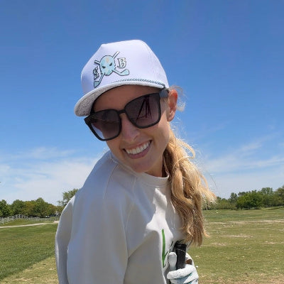 Woman on a golf course wearing a white cap, sunglasses, and holding a golf club.