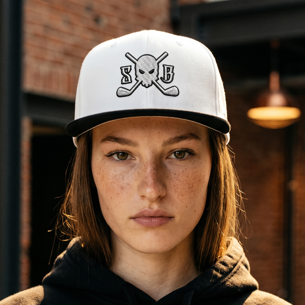 A person wearing a white baseball cap with a black brim and a black-and-white skull-and-crossbones graphic with the letters 'SB' on the front, paired with a black hoodie featuring a matching chest graphic. The background is a blurred brick alleyway.