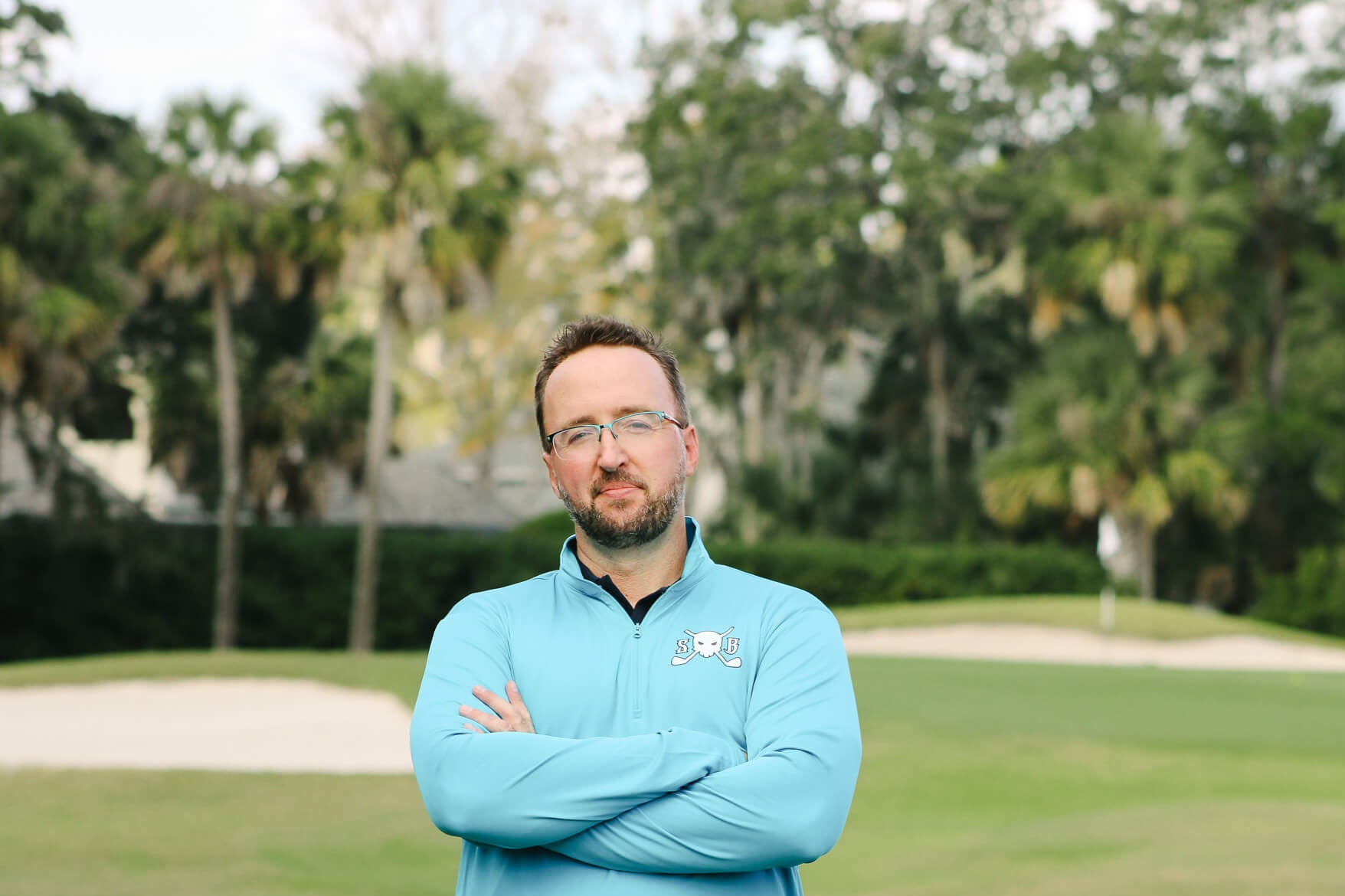 Man in a light blue jacket standing on a golf course with trees in the background