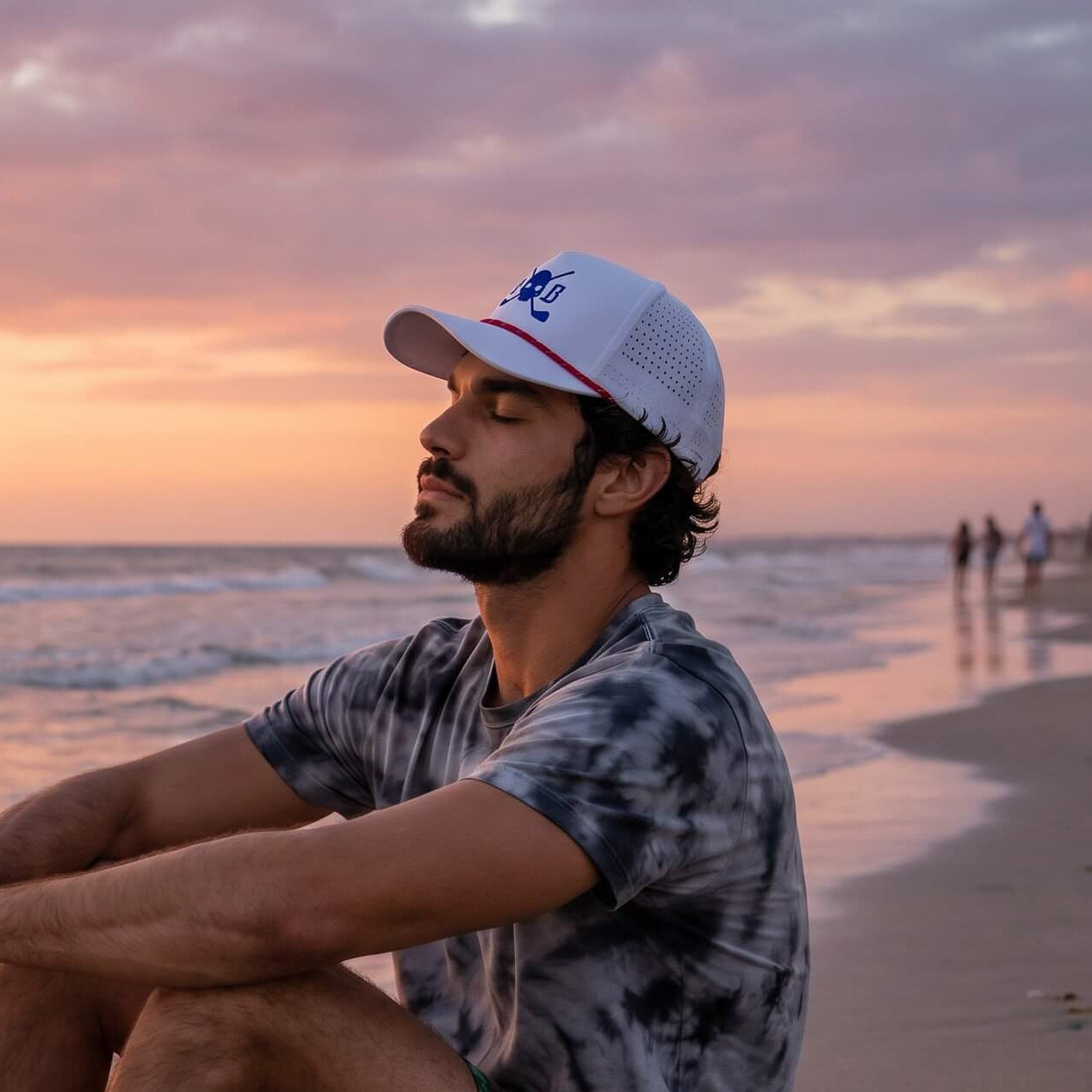 Man sitting on a beach at sunset wearing a tie-dye shirt and colorful shorts.