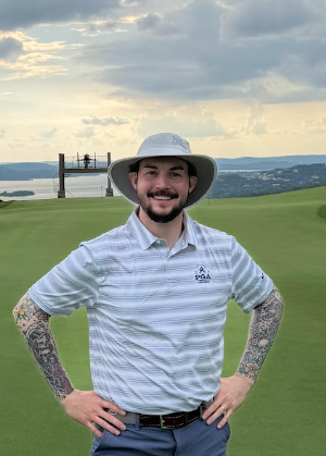 A man wearing a light blue and white striped polo shirt, slate blue shorts, and a wide-brimmed straw hat stands on a golf course green with a stone building and water in the background.