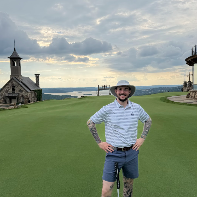A man wearing a light blue and white striped polo shirt, slate blue shorts, and a wide-brimmed straw hat stands on a golf course green with a stone building and water in the background.
