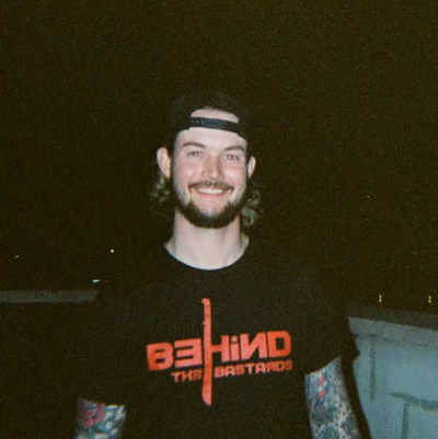 Man wearing a black t-shirt with 'BEHIND THE BASTARDS' text, standing against a dark background.