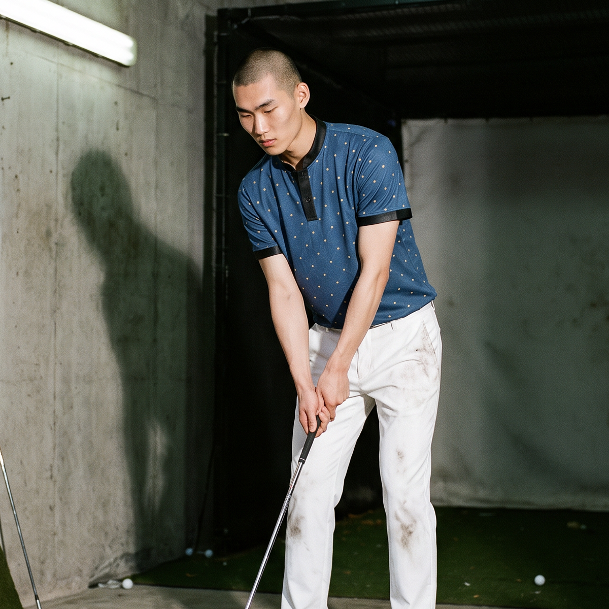 A man at an indoor golf driving range wearing a blue patterned polo shirt and white pants, preparing to hit a golf ball with a driver. Golf clubs are stored in the background, and a golf ball monitor is visible on the ground.