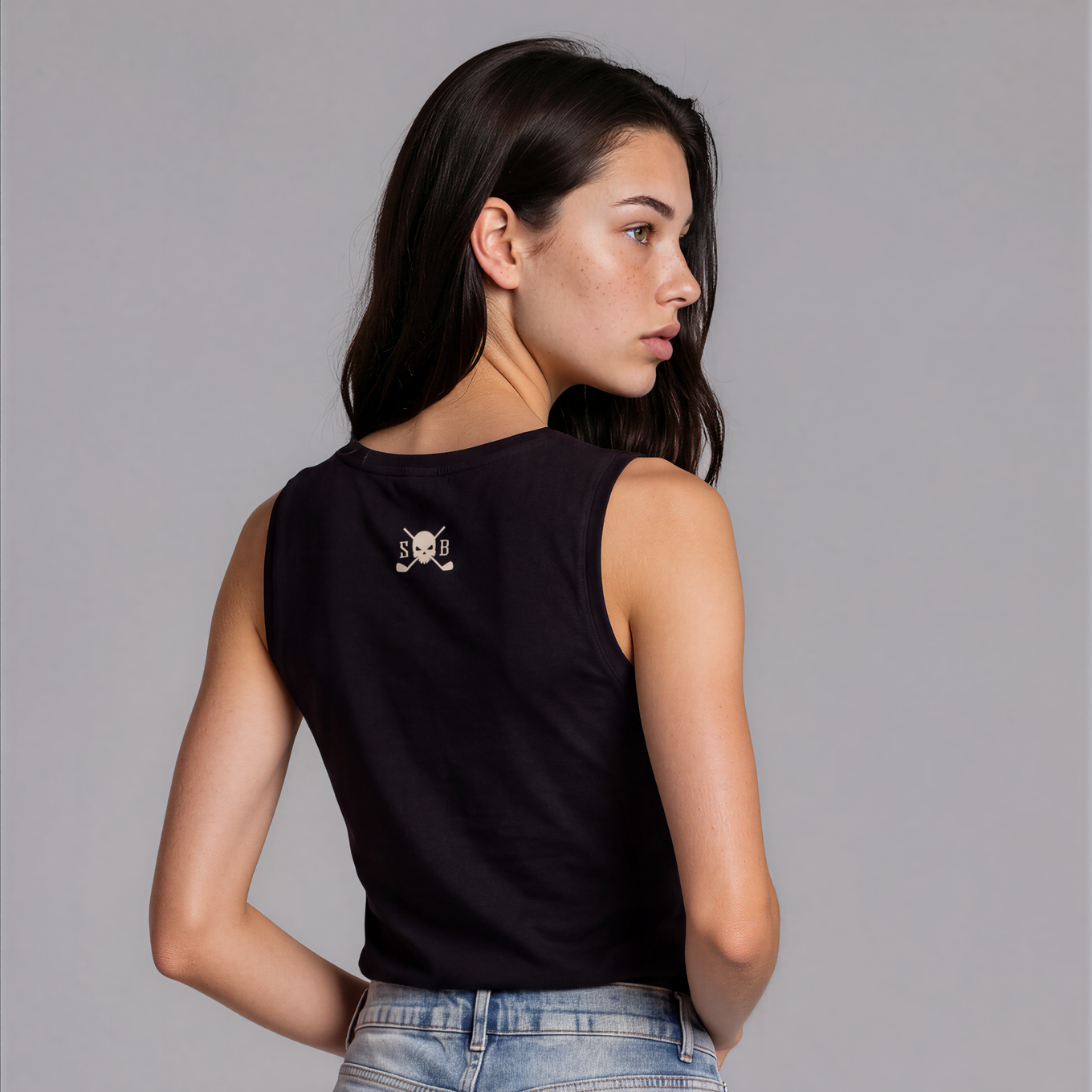 A woman shown from the back wearing a black sleeveless crop top with a small skull-and-crossbones graphic on the upper back, paired with light-wash denim shorts, against a plain gray studio background.