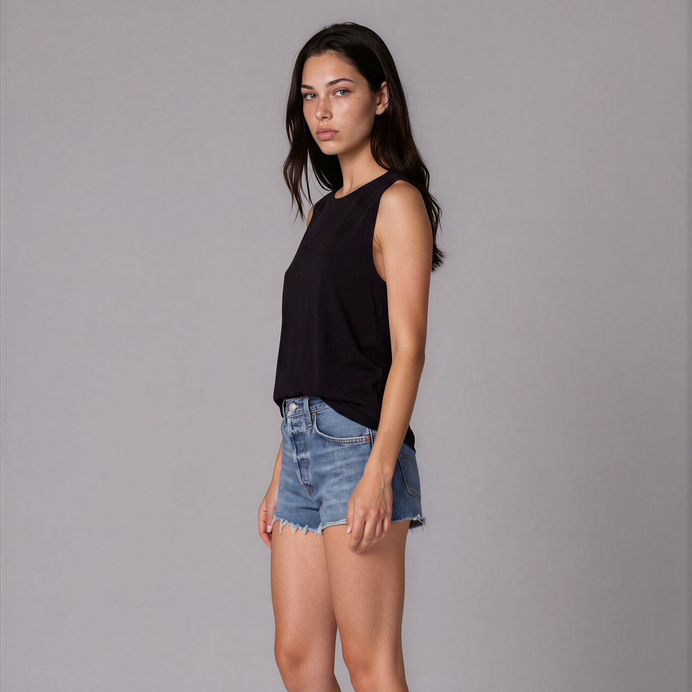 A woman stands in profile against a light gray studio background, wearing a sleeveless black top and light blue denim shorts. No visible product brand is shown; the small 'AI Generated | BOTIKA' text appears as a watermark/label rather than a product brand.