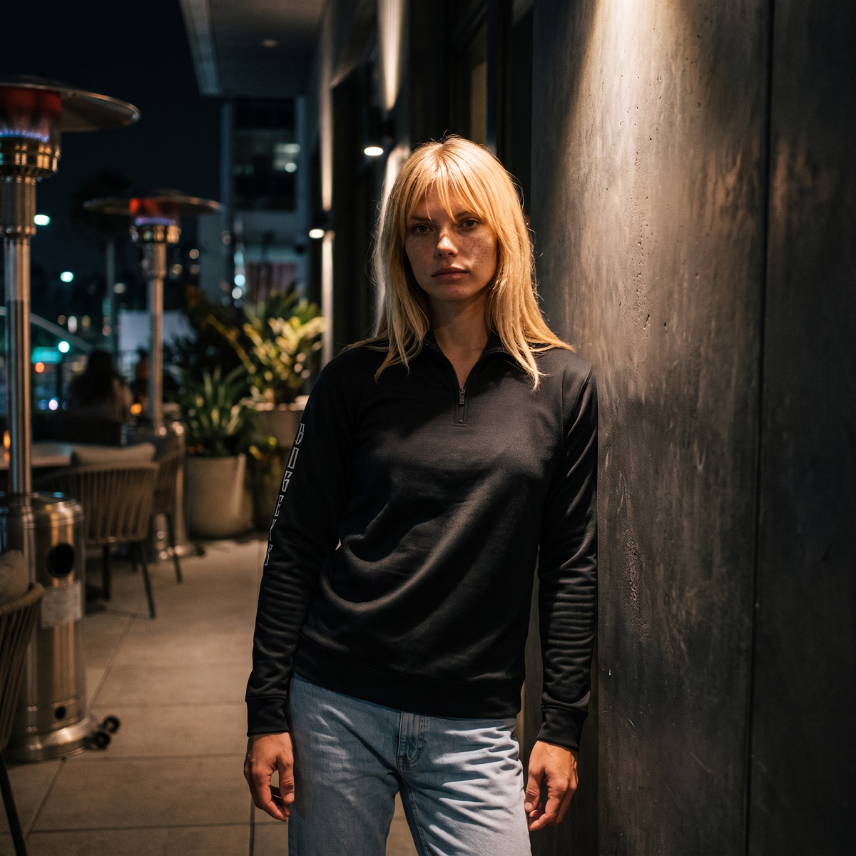 A woman with blonde hair stands against a dark wall at night, wearing a black long-sleeve quarter-zip pullover and light blue jeans.