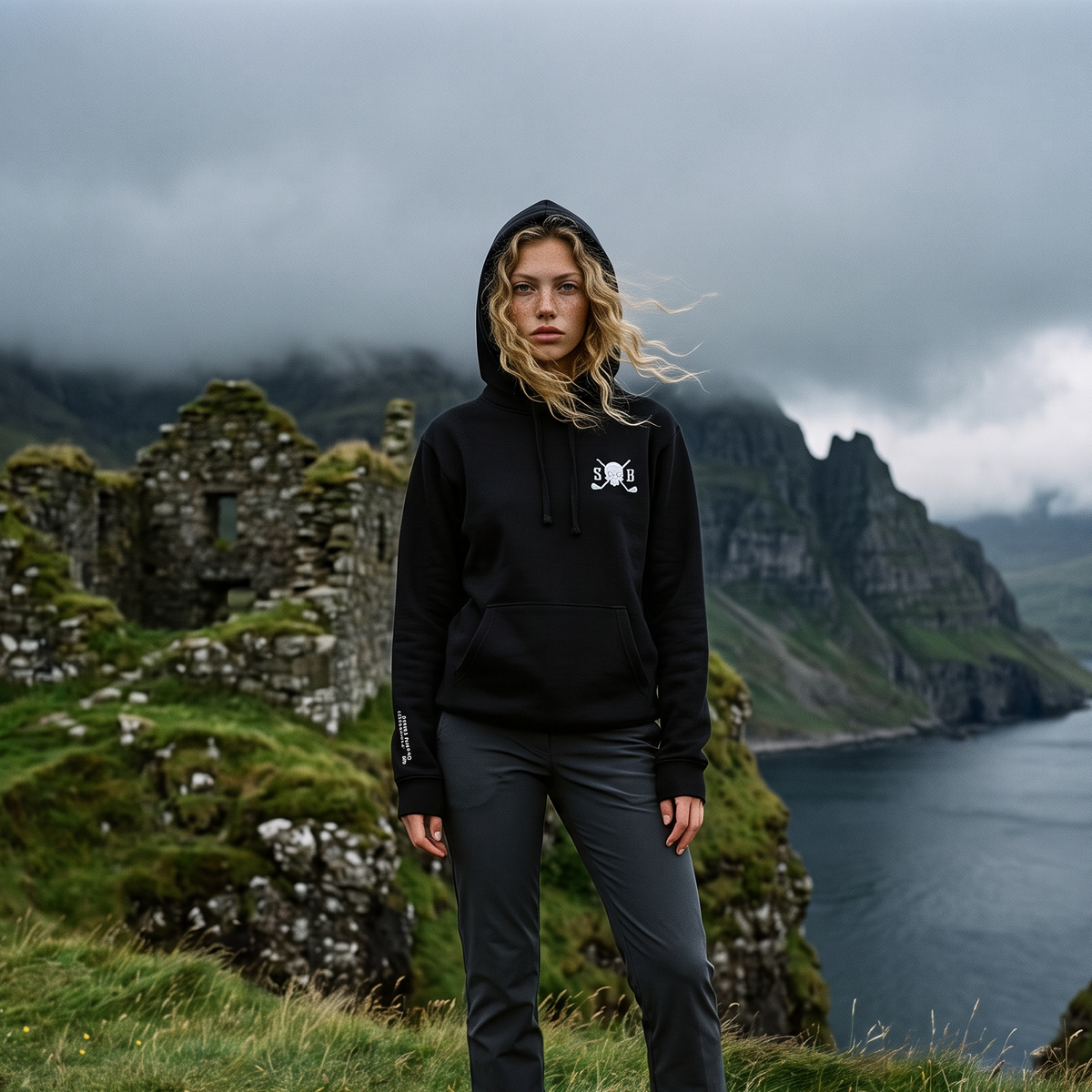 A woman stands outdoors on a grassy cliffside with stone ruins and a dramatic ocean landscape in the background, wearing a black pullover hoodie with a small white chest graphic and dark gray pants.