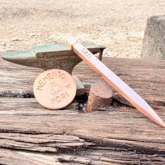 A copper-colored metal point tool, likely a chisel or engraving point, resting on a weathered wooden surface next to a bronze anvil and a copper coin for scale.