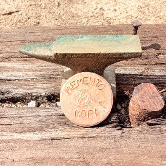A miniature anvil-shaped keepsake coin with a round bronze-colored center featuring the engraved words 'MEMENTO MORI' and a small green patina top, displayed on a weathered wooden surface outdoors.