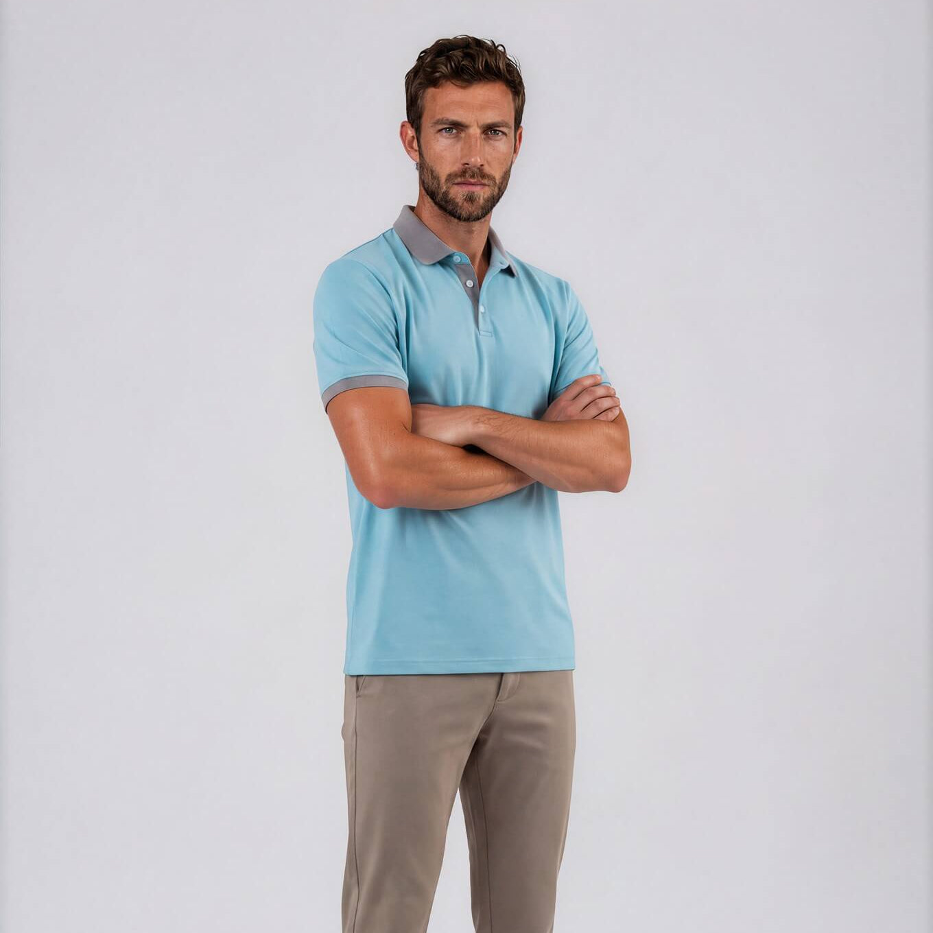 A man modeling a light blue Skull & Bogeys Golf Co. short-sleeve polo shirt with taupe pants and gray slip-on shoes, standing with arms crossed against a light gray studio background.