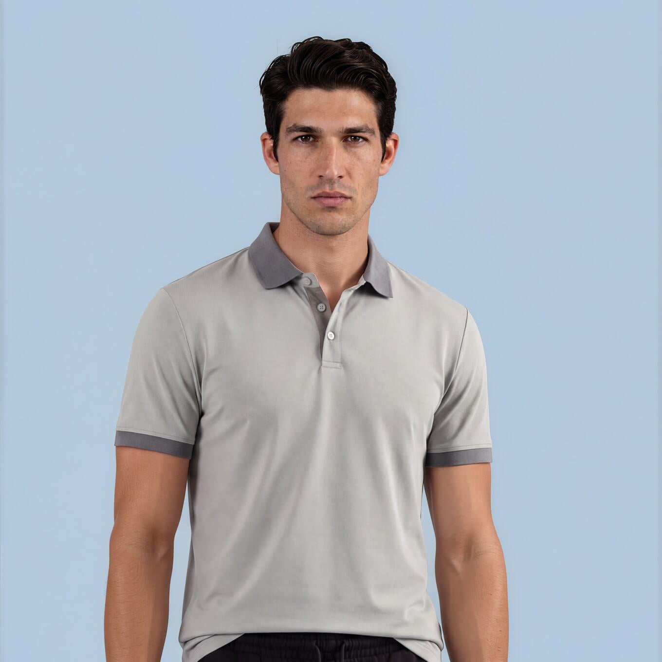 A man modeling a light gray short-sleeve polo shirt with dark gray trim on the collar and sleeve cuffs, paired with black shorts, against a solid light blue background.
