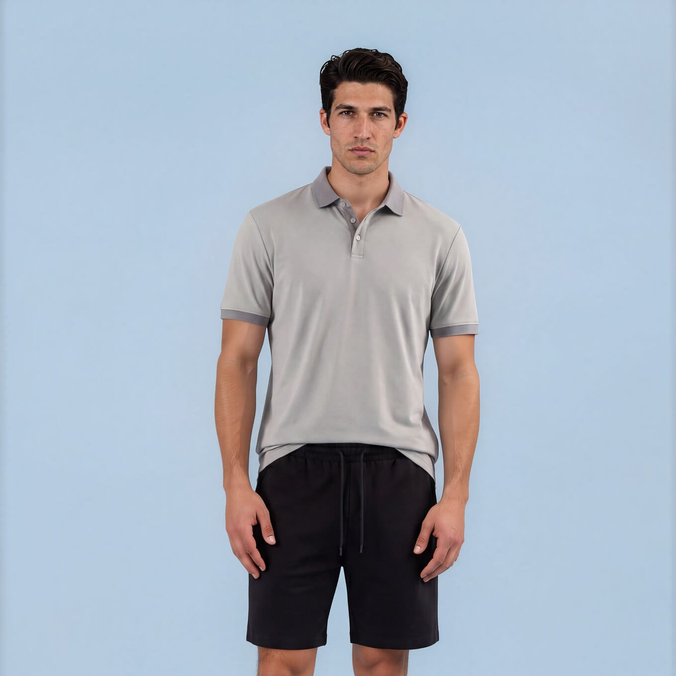A man modeling a light gray Skull & Bogeys Golf Co. short-sleeve polo shirt and black drawstring shorts against a solid light blue background.