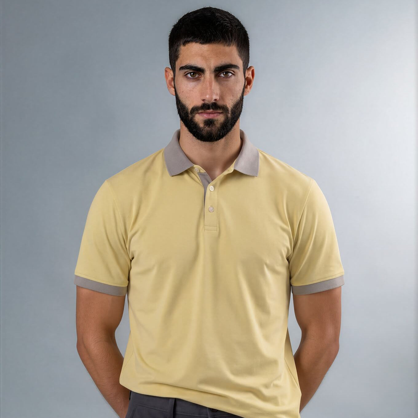 A man wearing a pale yellow Skull & Bogeys Golf Co. polo shirt with a contrasting gray collar and sleeve cuffs, paired with dark gray trousers, standing against a neutral gray studio background.