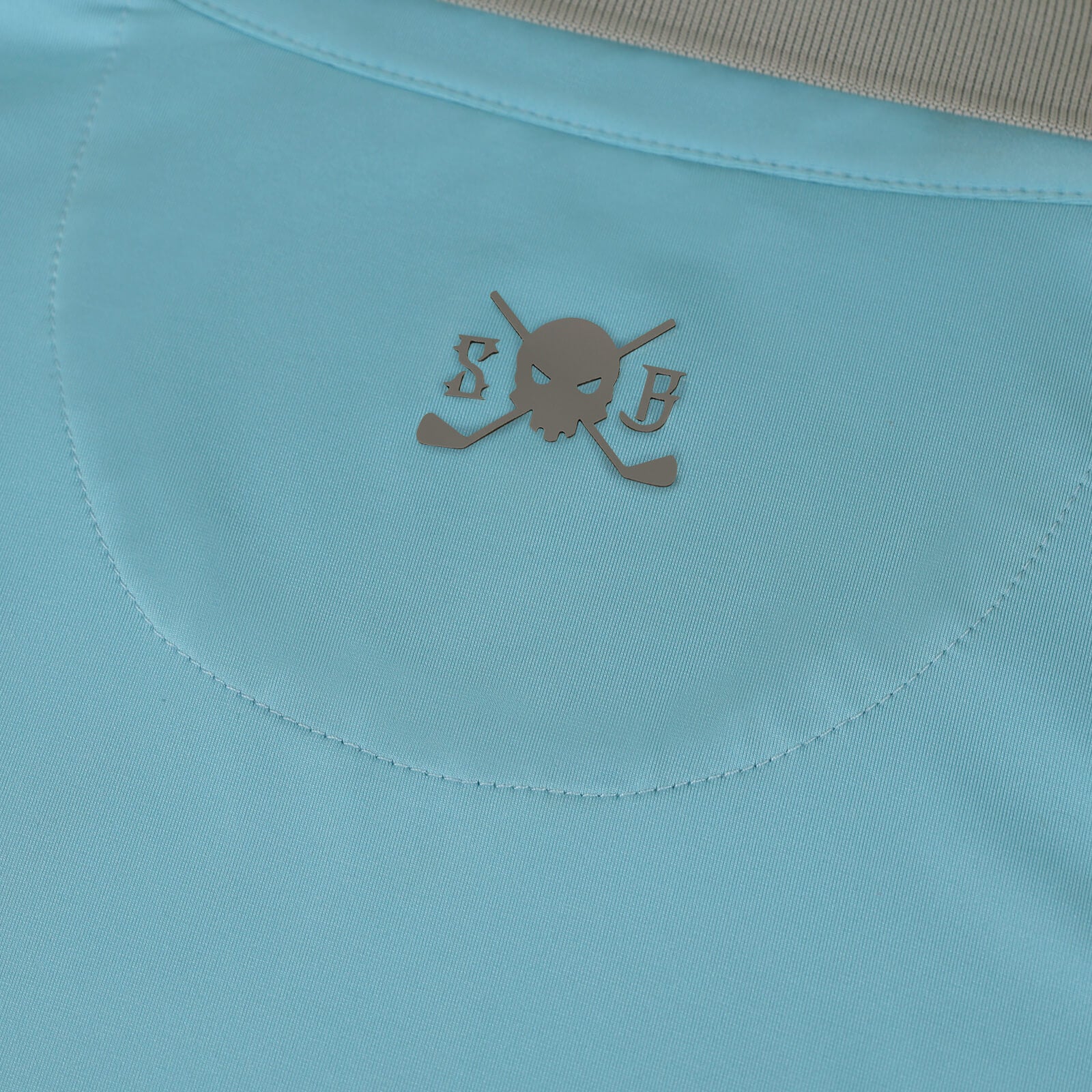 Close-up of a light blue Skull & Bogeys Golf Co. garment featuring a metallic bronze skull and crossed golf clubs logo with the letters 'S' and 'B'.