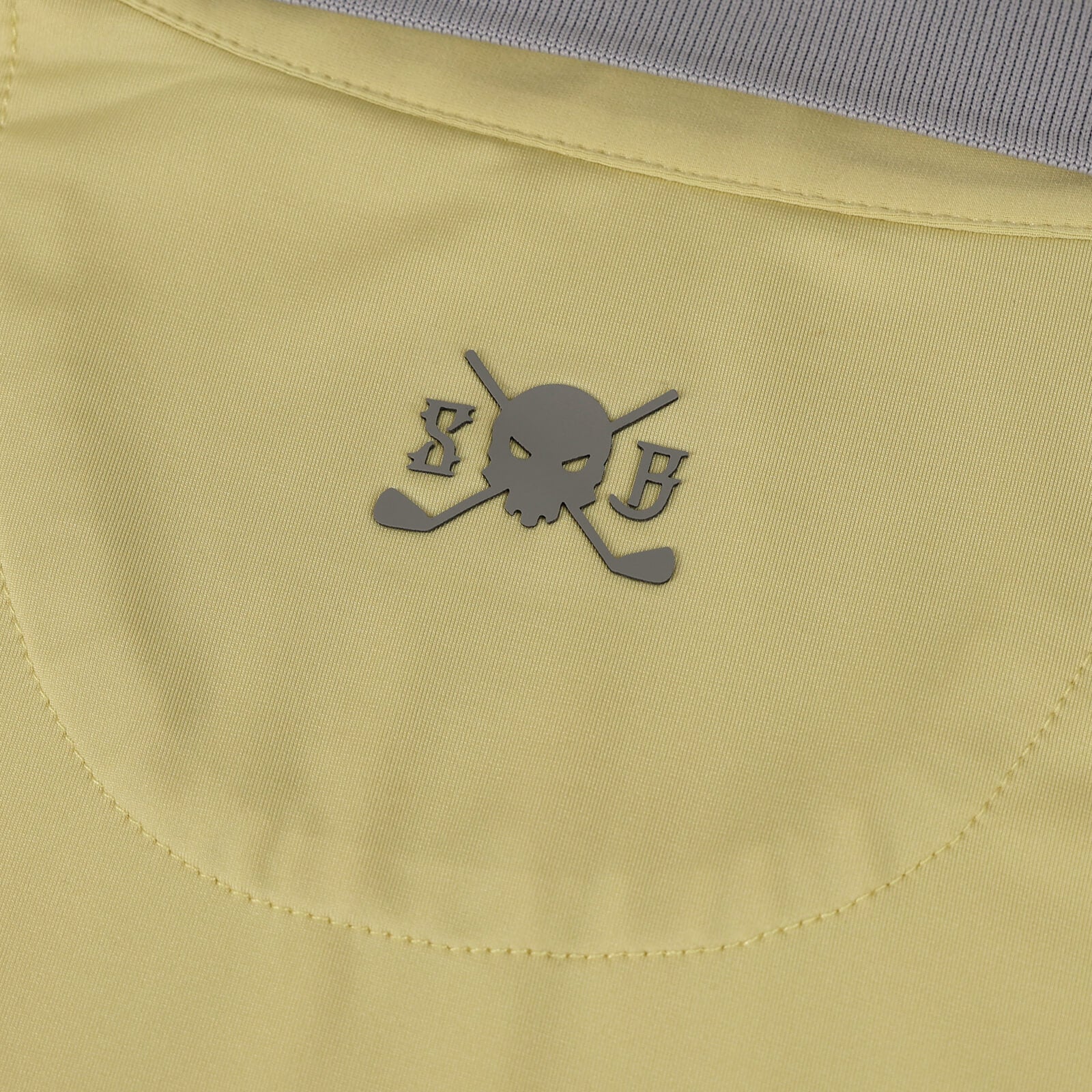 Close-up of a pale yellow Skull & Bogeys Golf Co. garment with a gray skull-and-crossbones logo featuring the letters S and B, plus a ribbed gray collar at the top.