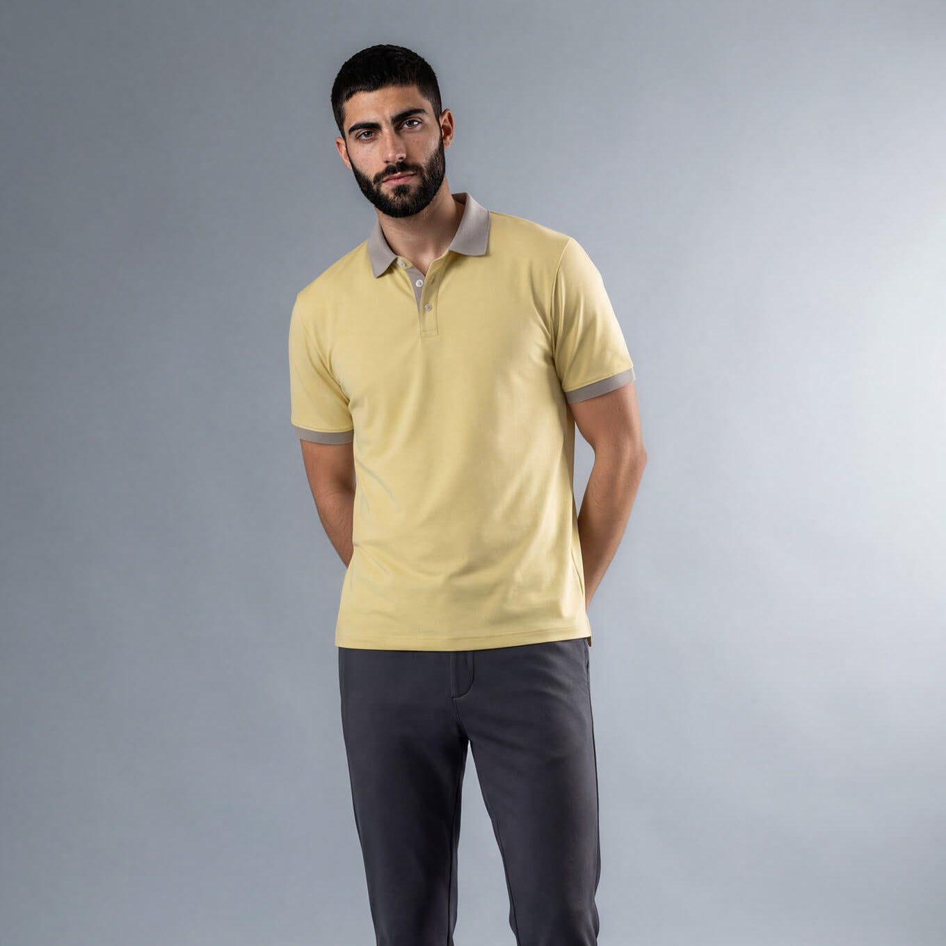 A man wearing a pale yellow short-sleeve polo shirt with a gray collar and sleeve trim, paired with dark gray trousers and black shoes, standing against a gray studio background.