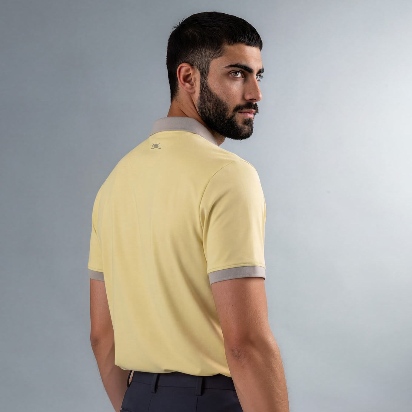A man wearing a pale yellow Skull & Bogeys short-sleeve polo shirt with gray trim on the collar and sleeve cuffs, paired with navy trousers, shown from a three-quarters back view against a neutral gray studio background.