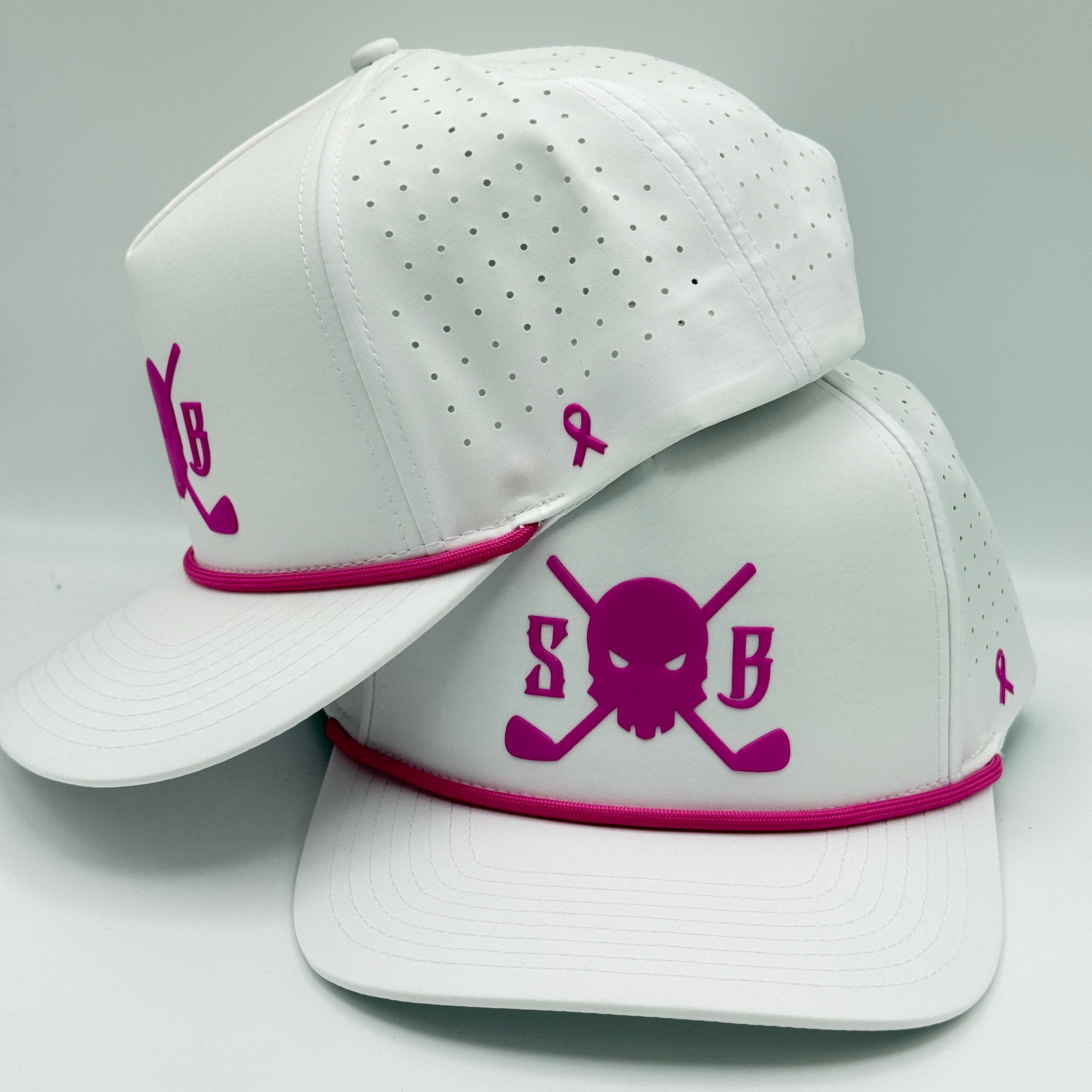 Two white baseball caps with pink accents, stacked together on a light gray background. The front cap features a pink skull and crossbones design with the letters 'S' and 'B', and a pink awareness ribbon on the side.