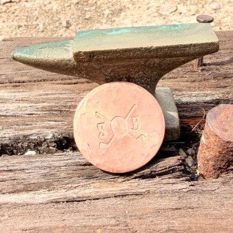 A small anvil-shaped object with a greenish patina sits on a weathered wooden surface, with a round copper-colored coin placed in front of it for scale.