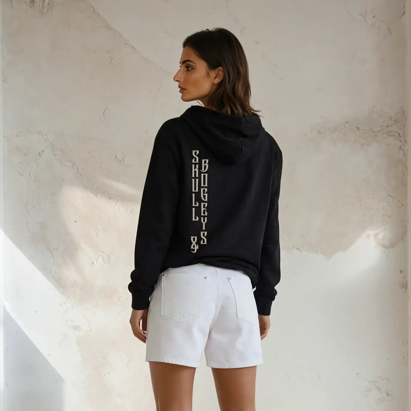 Woman wearing a black hoodie with text on the back and white shorts, standing against a light-colored wall.