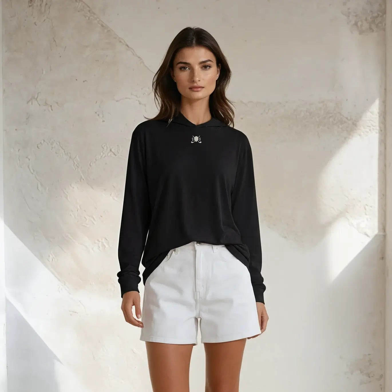 Woman wearing a black sweater and white shorts standing against a textured white wall.