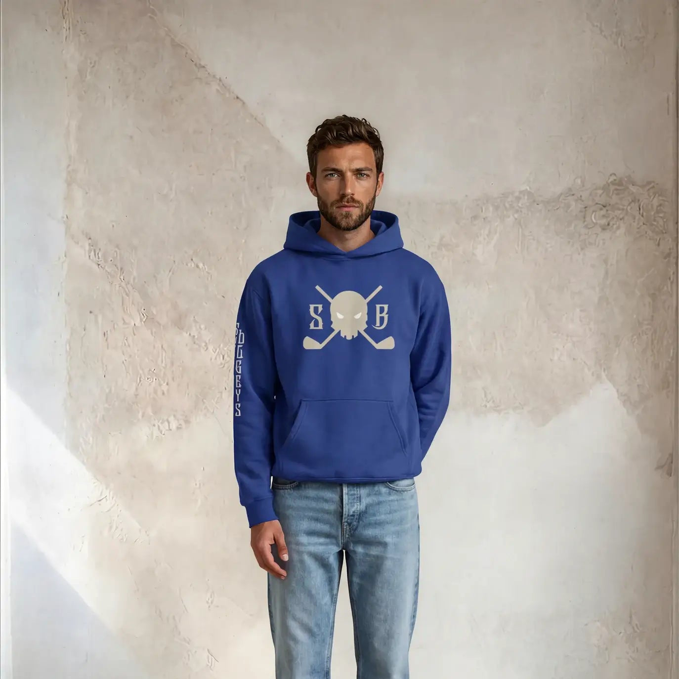 Man wearing a blue hoodie with a logo and light blue jeans standing against a textured wall.