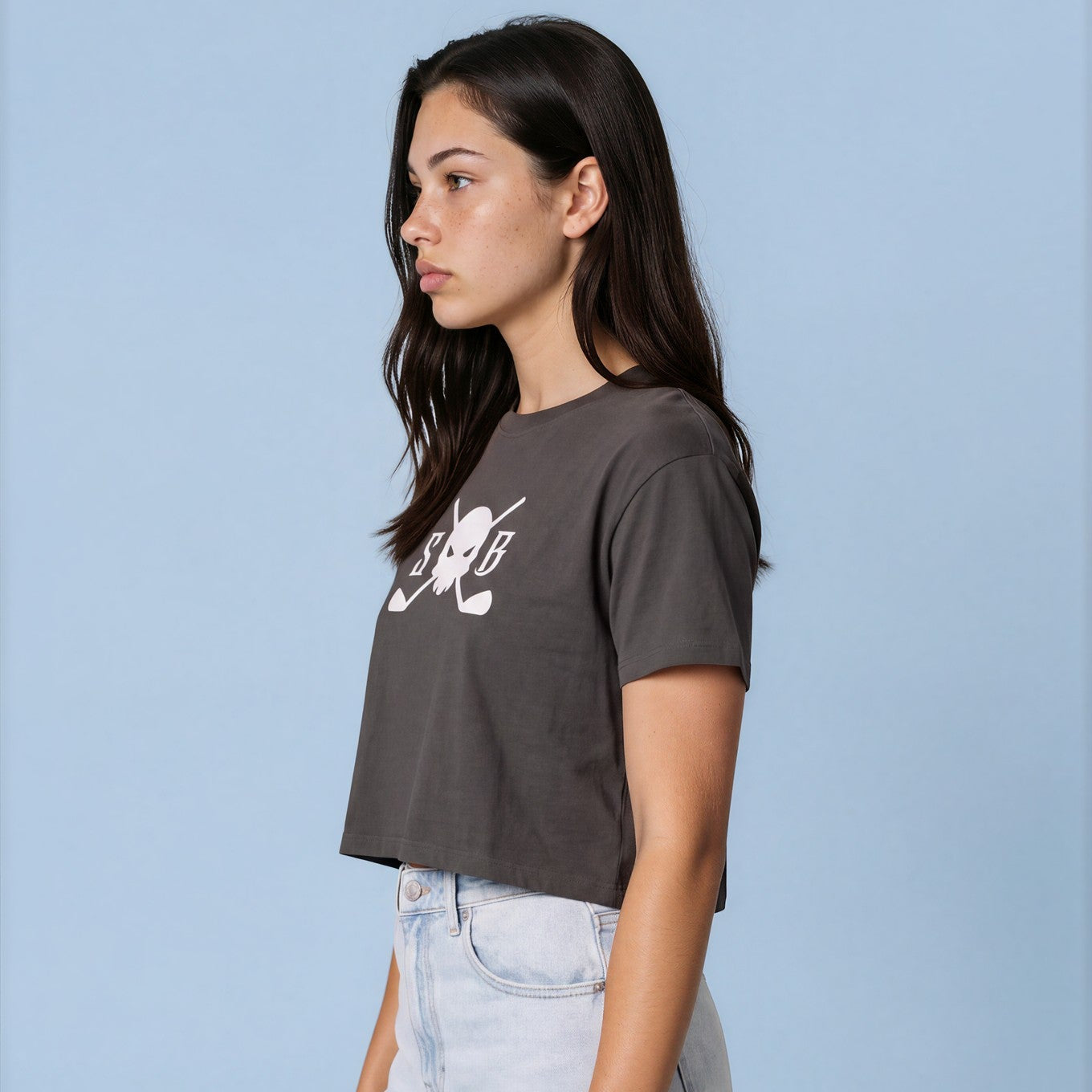 A woman wearing a dark gray cropped t-shirt with a white graphic print and light wash wide-leg jeans, standing against a solid light blue background.