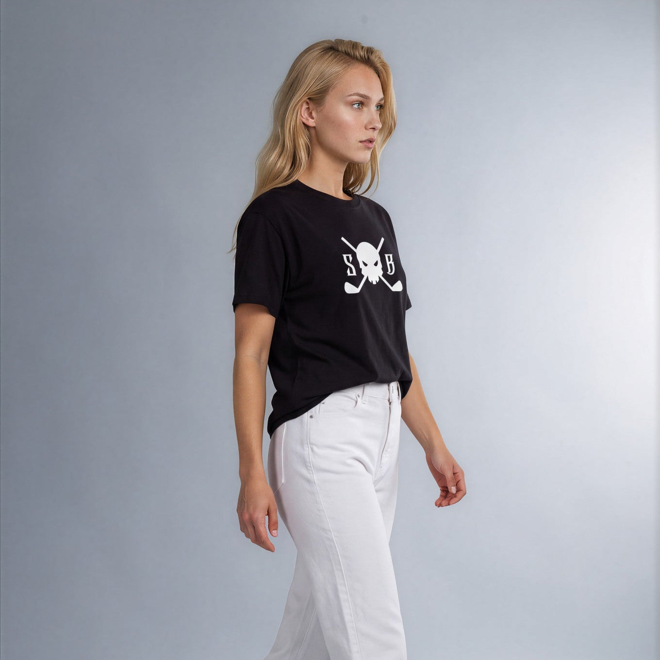 A young woman walking in profile wearing a black short-sleeve t-shirt with a white skull and crossbones graphic, white pants, and black strappy sandals, set against a light gray studio background.