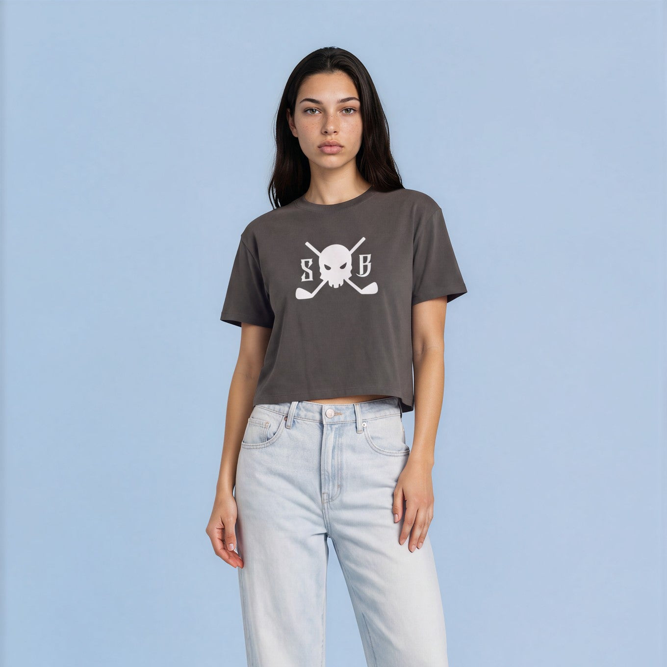 A woman modeling a dark gray cropped t-shirt with a white 'SMB' and crossed golf clubs graphic, paired with light-wash wide-leg jeans and white sneakers, against a solid light blue background.