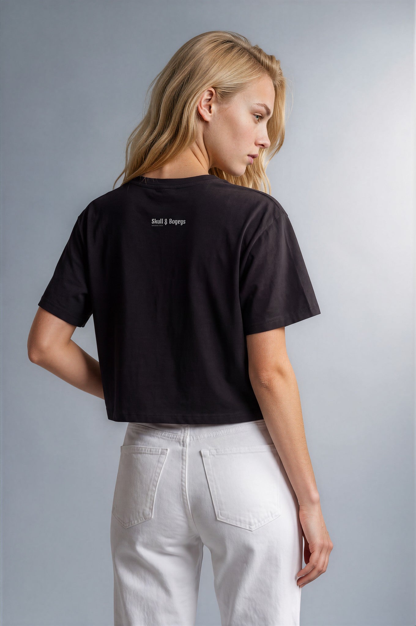 A woman shown from the back wearing a black short-sleeve crop top with small white text on the upper back and white pants, photographed against a plain light gray studio background.