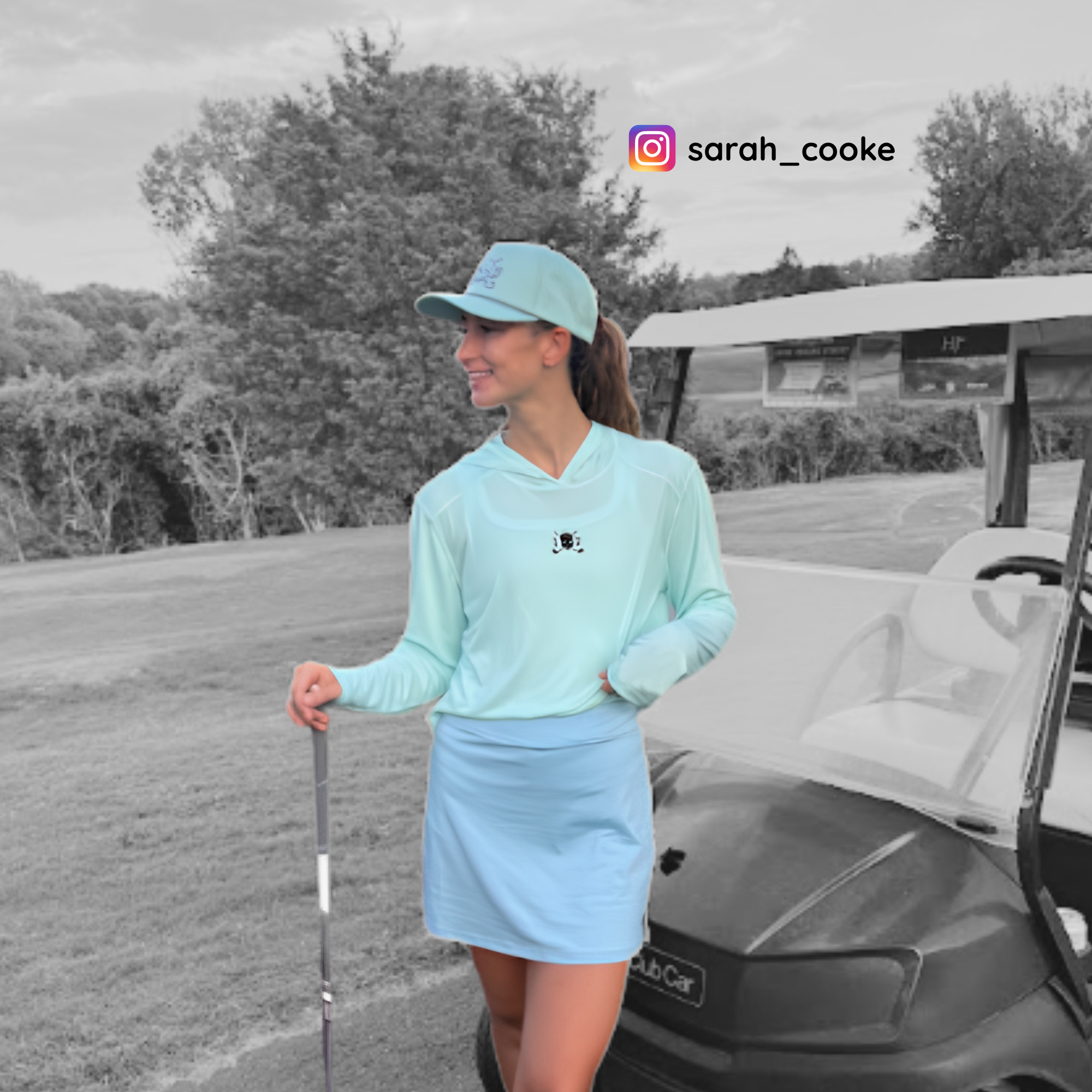A woman stands on a golf course next to a golf cart, wearing a light aqua long-sleeve hooded top, a matching light blue skirt, and a light blue cap. The background is desaturated in grayscale, with trees and grass visible.