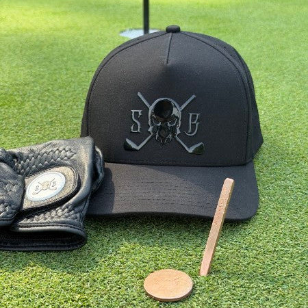 A black baseball cap with a skull and crossed golf clubs logo sits on a green grass surface, alongside a black golf glove, a wooden tee, and a gold coin. The cap features a tonal black embroidered logo with the letters 'SG'.