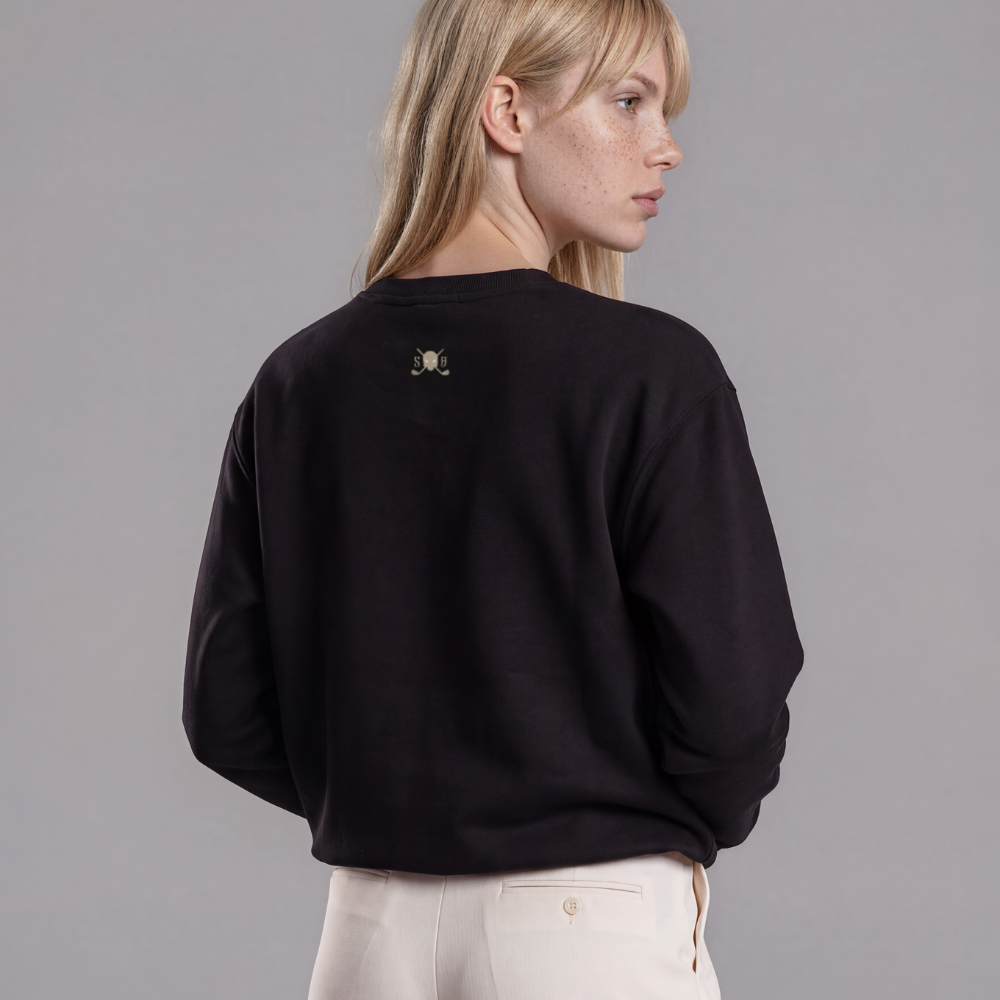 A woman shown from the back wearing a black crewneck sweatshirt with a small gold Botika logo on the upper back, paired with cream-colored wide-leg trousers, against a solid gray studio background.