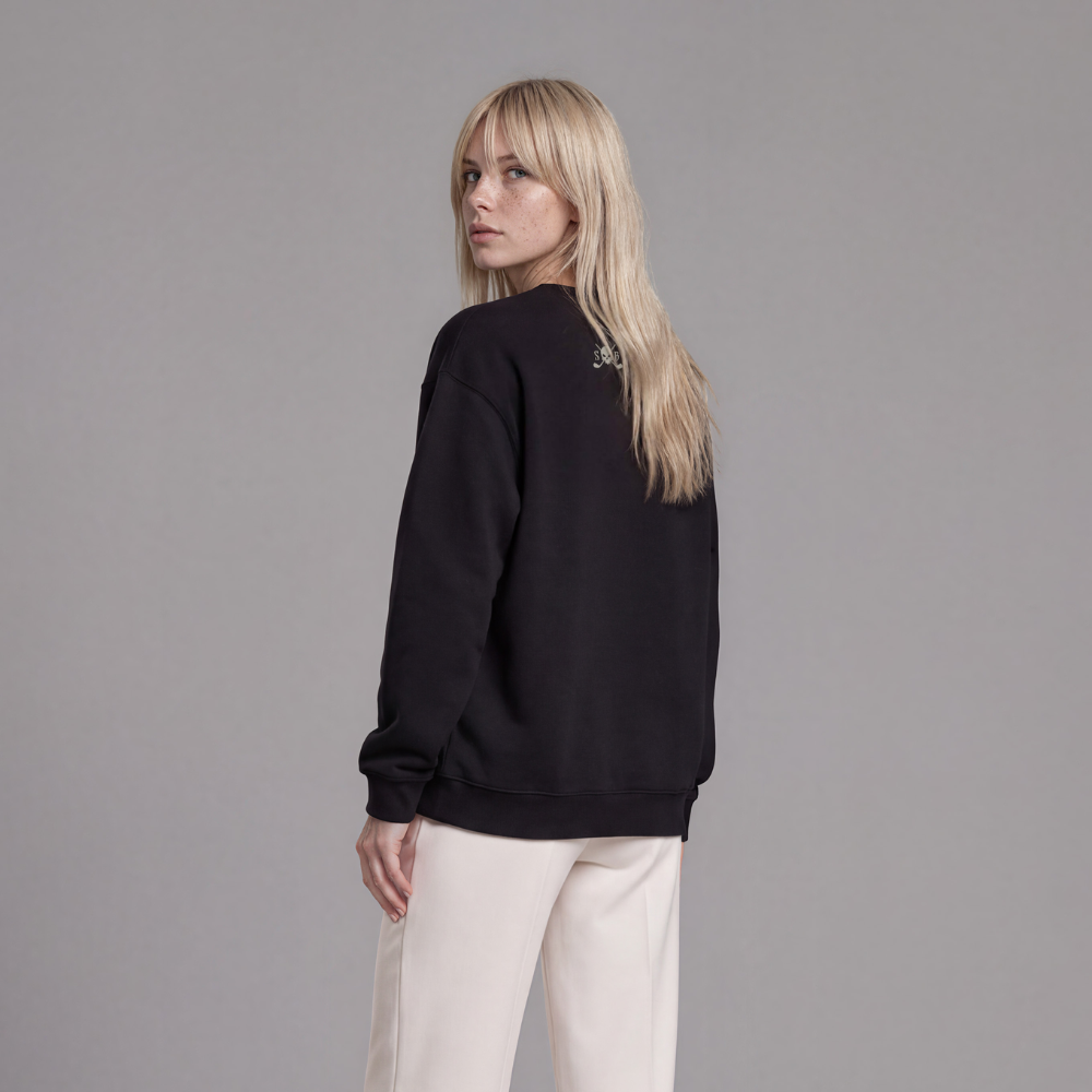 A woman stands in a studio wearing a black crewneck sweatshirt, off-white wide-leg trousers, and black platform shoes, photographed from the back against a light gray background.
