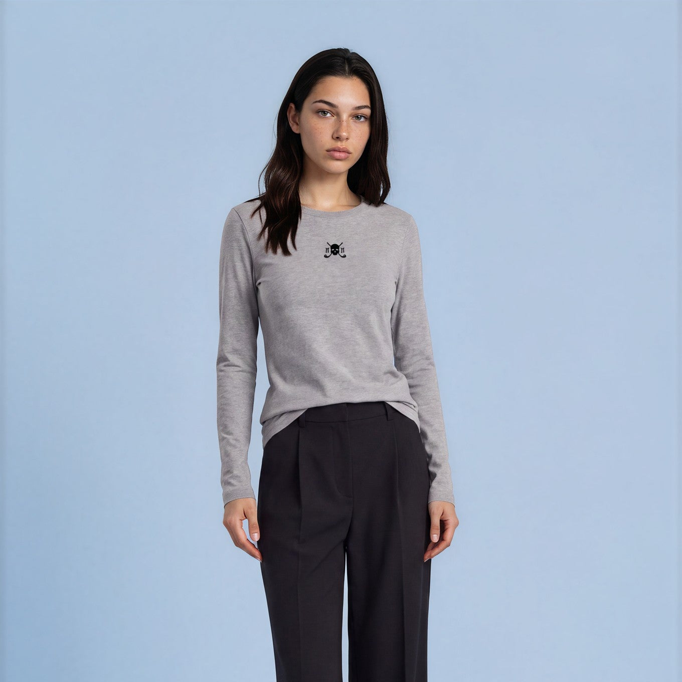 A woman stands against a light blue studio background wearing a light gray long-sleeve top with a small black chest logo and black wide-leg trousers.