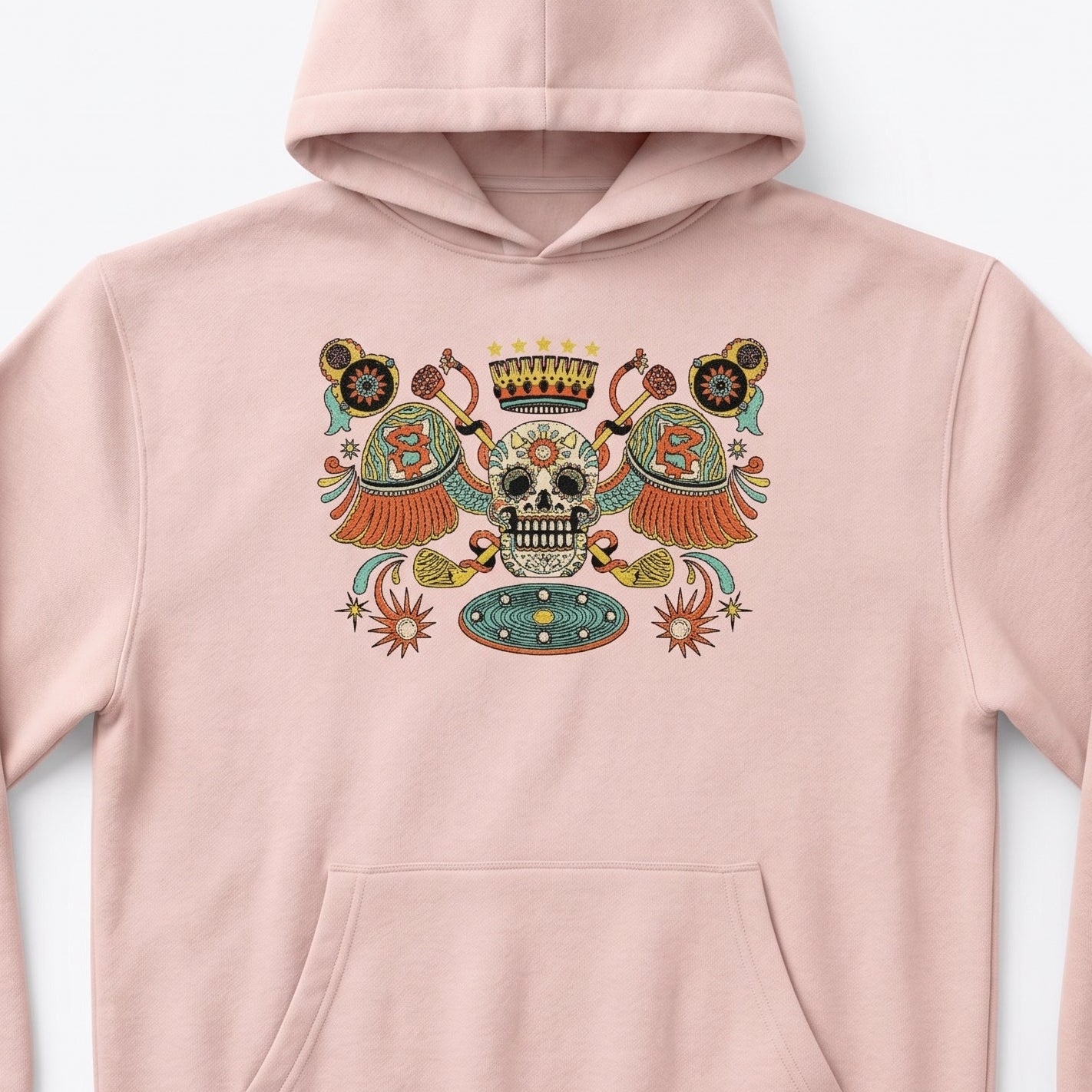 A light pink pullover hoodie with a colorful Day of the Dead (Dia de los Muertos) sugar skull graphic on the chest, displayed against a clean white background.