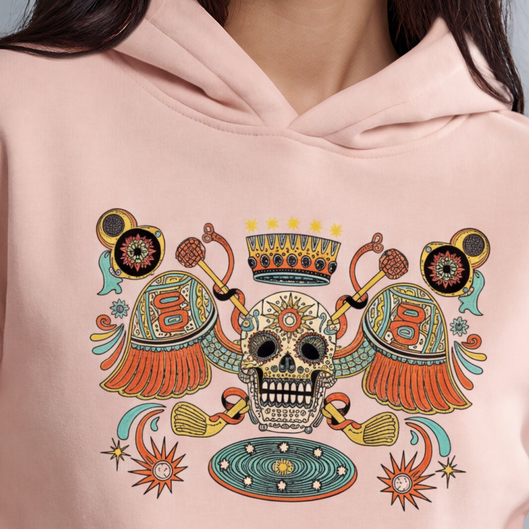 A woman modeling a light pink hoodie with a colorful Day of the Dead (Dia de los Muertos) style graphic on the chest, paired with black jeans, against a solid gray studio background.