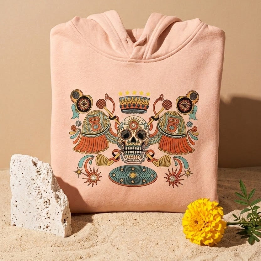 A light pink hoodie with a colorful Day of the Dead (Dia de los Muertos) sugar skull graphic, styled on a sandy surface with a stone and a yellow flower against a beige background.