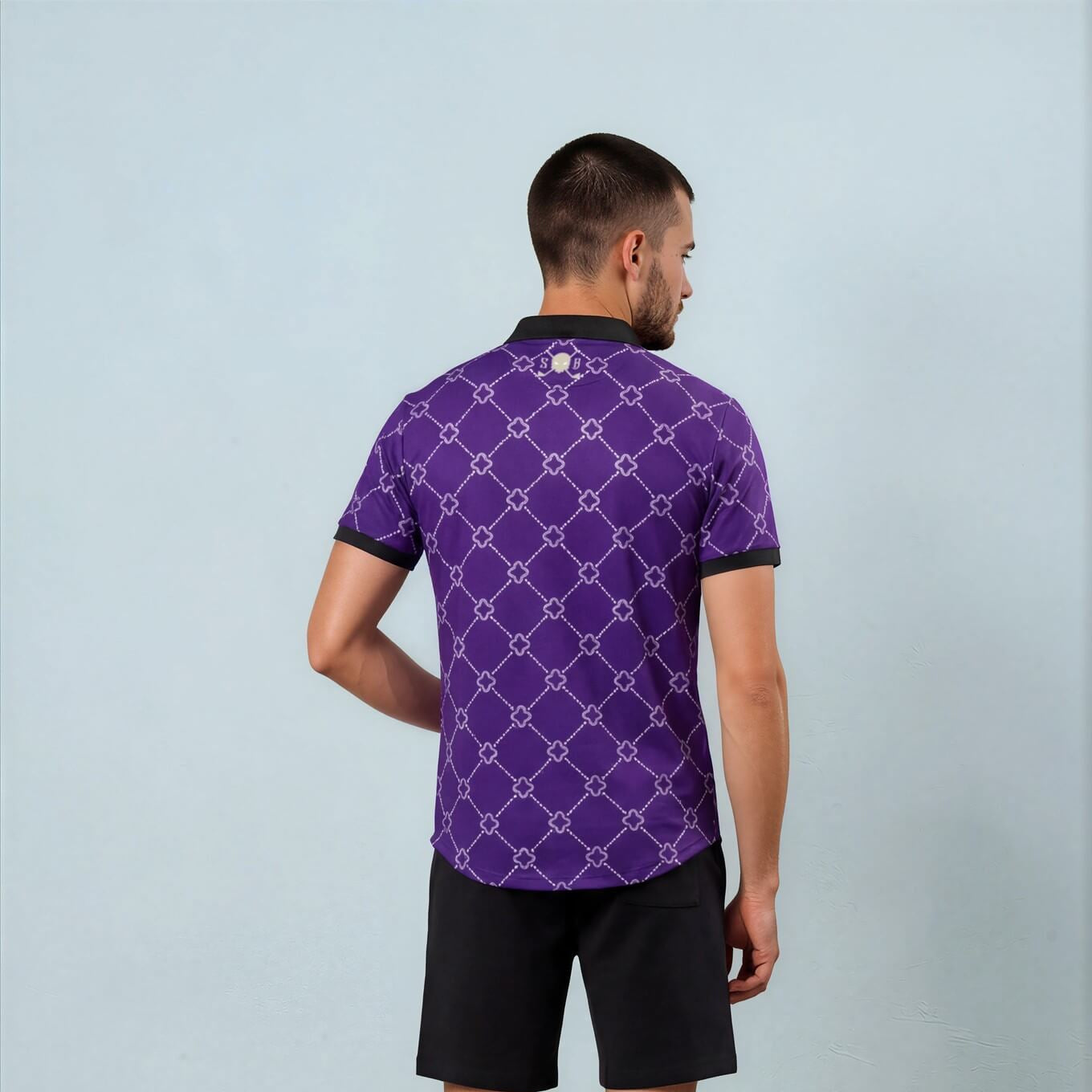 Man wearing a purple patterned polo shirt and black shorts against a light blue background