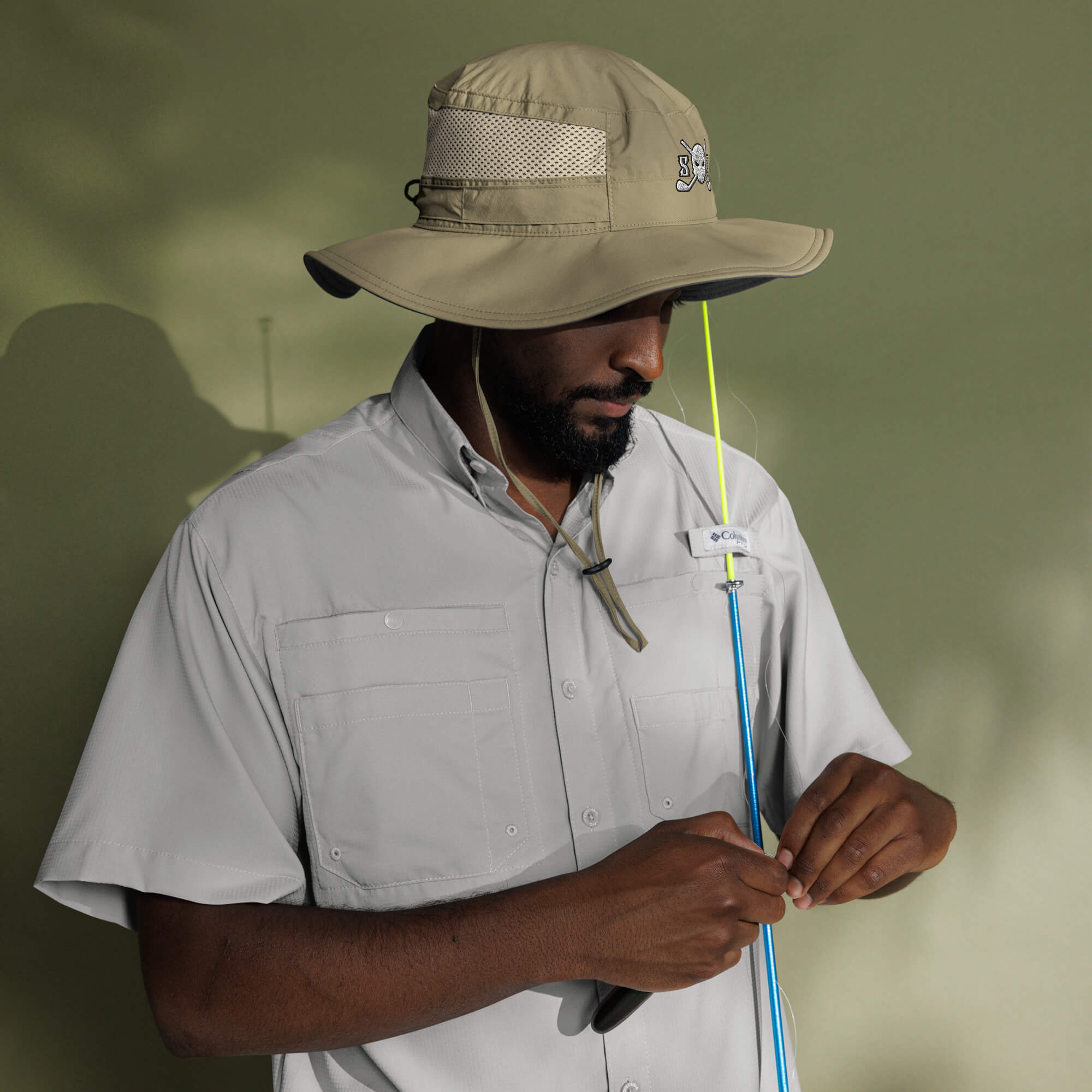 Man wearing a Skull & Bogeys Golf Co. beige hat and shirt, holding a fishing rod against a blurred green background